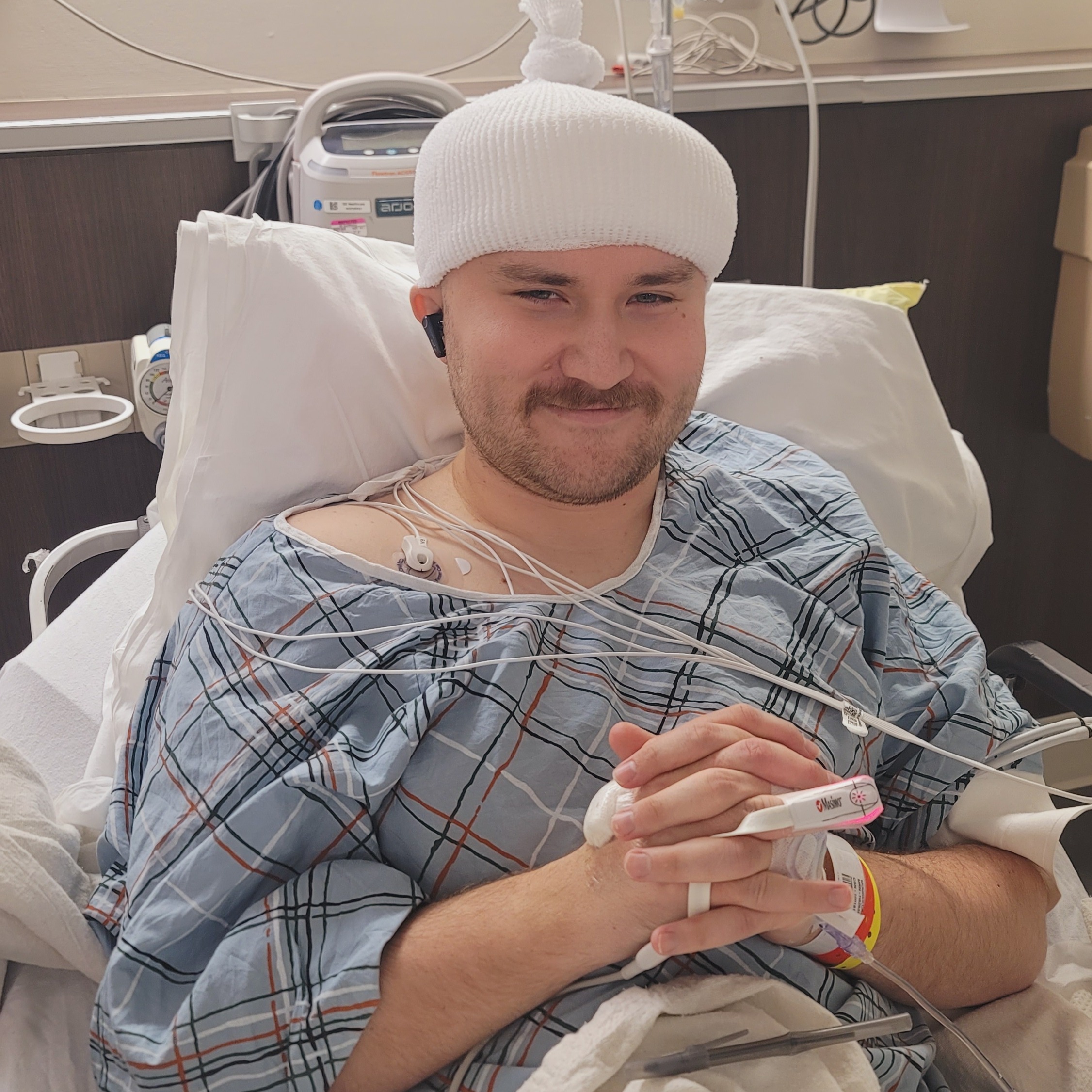 Caleb in a hospital bed after surgery, wearing a post-operative cap, with monitors nearby.